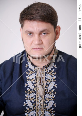 stern look of blue eyes handsome man in Ukrainian embroidered shirt looks straight into frame he has light beard compressed lips thick hair a blue shirt and embroidery on a white background close-up 112204600