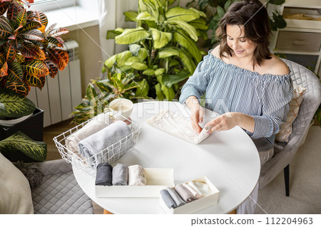 Smiling plus size woman folding linen nightdress in PVC container metal basket storage organizing 112204963