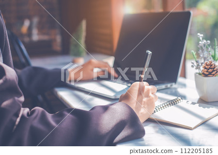 Business woman hand writing on a notepad with a pen and use a laptop computer in the office. 112205185
