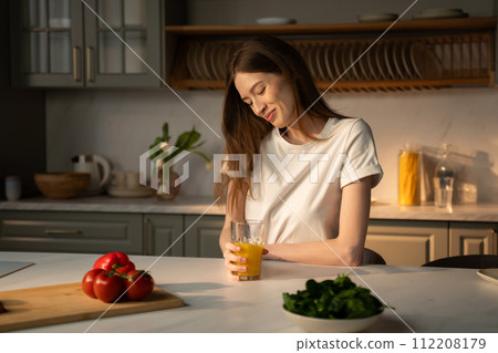 Young Woman Enjoying a Fresh Glass of Orange Juice in a Sunny Kitchen 112208179