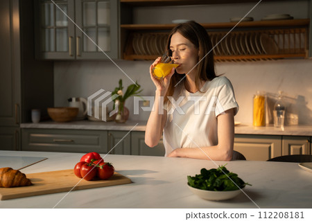Young Woman Enjoying a Fresh Glass of Orange Juice in a Sunny Kitchen Young Woman Enjoying a Fresh Glass of Orange Juice in a Sunny Kitchen 112208181
