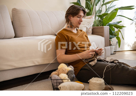 Feminine hipster man crochet handmade use hook knitting needles and wool yarn relaxing on couch Feminine hipster man crochet handmade use hook knitting needles and wool yarn relaxing on couch 112209023