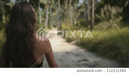 Young hiker woman with long brown hair, back view, walking in jungle forest. Girl hiking alone. Tourist on jungle park trail, looking around. Exotic travel, tourism, holiday, hiking, active lifestyle. 112212368