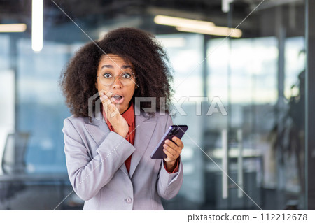 Portrait of shocked young African American woman in business suit standing in office, holding mobile phone, looking at camera in surprise, holding hand to face. Portrait of shocked young African American woman in business suit standing in office, holding mobile phone, looking at camera in surprise, holding hand to face. 112212678
