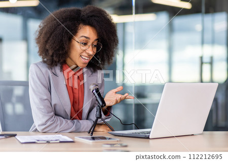 African american young woman sitting in the office at the desk in a suit and talking through a microphone on a video call on a laptop, smiling and gesturing with her hands. African american young woman sitting in the office at the desk in a suit and talking through a microphone on a video call on a laptop, smiling and gesturing with her hands. 112212695