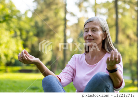 Close-up photo of senior gray-haired woman doing yoga outside in park, sitting in lotus position with closed eyes and meditating. Close-up photo of senior gray-haired woman doing yoga outside in park, sitting in lotus position with closed eyes and meditating. 112212712