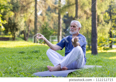 Senior gray-haired man doing yoga in the park, sitting on a mat in the lotus position and meditating with closed eyes alone. 112212715