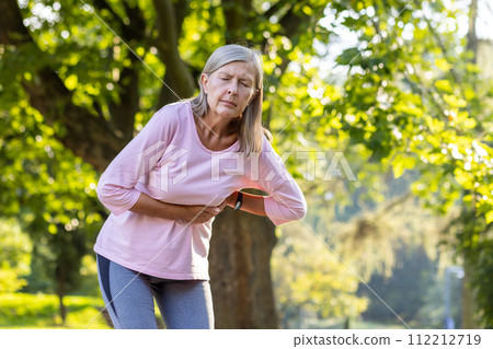 An older gray-haired woman is standing in the park bent over and holding her hands to the side of her body, feeling severe pain, needing medical help and rest. An older gray-haired woman is standing in the park bent over and holding her hands to the side of her body, feeling severe pain, needing medical help and rest. 112212719