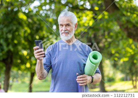 Portrait of a smiling and healthy gray-haired man standing in the park, holding a phone, a yoga mat and a bottle of water, confidently looking at the camera. Portrait of a smiling and healthy gray-haired man standing in the park, holding a phone, a yoga mat and a bottle of water, confidently looking at the camera. 112212731