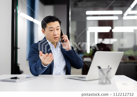 Serious young Asian man talking on mobile phone, sitting in office at desk and working on laptop and documents. Serious young Asian man talking on mobile phone, sitting in office at desk and working on laptop and documents. 112212732