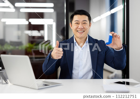 Portrait of a smiling young Asian man in a business suit sitting in the office, holding an asthma and allergy inhaler in his hand, and showing the camera a super gesture with his finger. Portrait of a smiling young Asian man in a business suit sitting in the office, holding an asthma and allergy inhaler in his hand, and showing the camera a super gesture with his finger. 112212751
