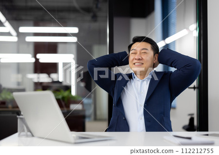 Satisfied with the result, a young Asian male businessman sits relaxed in a chair at a desk in the office, hands behind his head, closes his eyes and rests with a smile. Satisfied with the result, a young Asian male businessman sits relaxed in a chair at a desk in the office, hands behind his head, closes his eyes and rests with a smile. 112212755