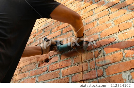 cutting the outline of a window in a brick wall with a grinder, a man in a black T-shirt and gloves cuts the masonry of the street wall of a building using a miter saw 112214162