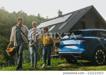 Family with electric car in front house with solar panels on roof. Solar, green energy and sustainable lifestyle of young family. 112215423