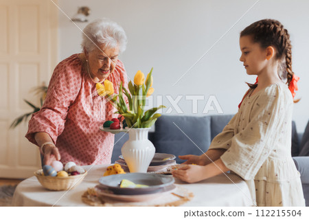 Grandmother with granddaughter setting table for traditional easter lunch. Recreating family traditions and customs. Grandmother with granddaughter setting table for traditional easter lunch. Recreating family traditions and customs. 112215504