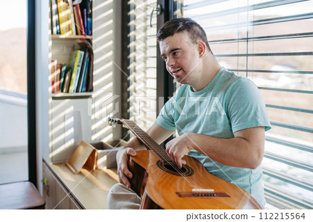 Young man with down syndrome playing acoustic guitar, sitting by window, holding and strumming guitar, making music. Young man with down syndrome playing acoustic guitar, sitting by window, holding and strumming guitar, making music. 112215564
