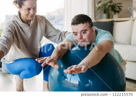 Young man with Down syndrome exercising at home with his mother on fitness ball. Workout routine for disabled man. 112215570