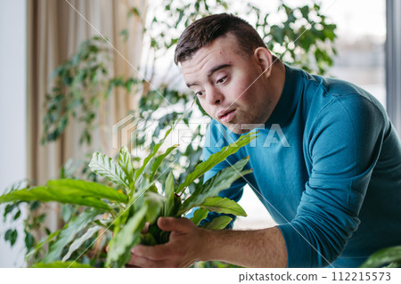 Young man with Down syndrome taking care of indoor plant, touching, snuggling plant leaf. Young man with Down syndrome taking care of indoor plant, touching, snuggling plant leaf. 112215573