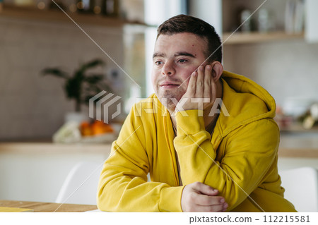 Young man with Down syndrome in kitchen, propping head up with his hand. Young man with Down syndrome in kitchen, propping head up with his hand. 112215581