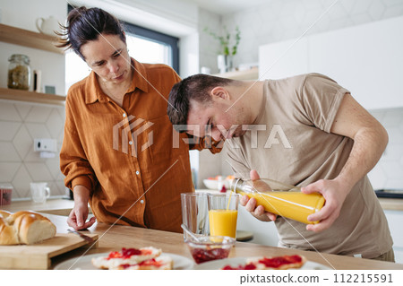 Young man with Down syndrome, pouring juice, preparing breakfast with his mother at home. Morning routine for man with Down syndrome. Young man with Down syndrome, pouring juice, preparing breakfast with his mother at home. Morning routine for man with Down syndrome. 112215591