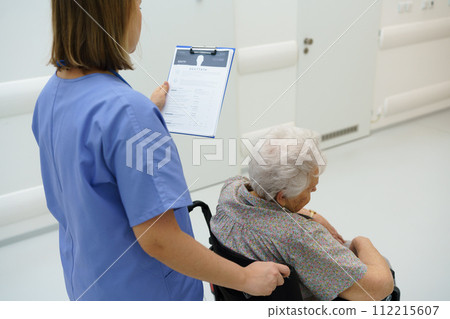 Nurse pushing senior patient in a wheelchair, reading medical document in clipboard. Emotional support for elderly woman. 112215607