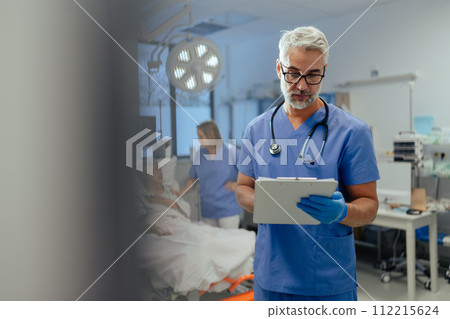 Portrait of handsome male doctor, patient in hospital bed behind. ER doctor examining senior patient, reading her medical test, lab results in clipboard. 112215624