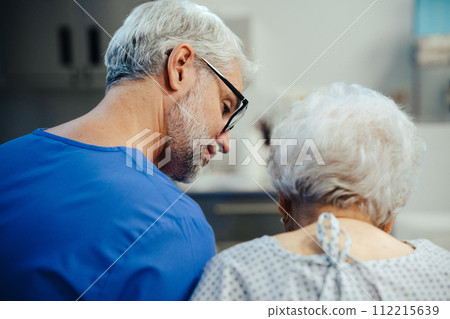 Supportive doctor soothing worried senior patient, discussing test result in emergency room. Compassionate emotional support for elderly patient. Supportive doctor soothing worried senior patient, discussing test result in emergency room. Compassionate emotional support for elderly patient. 112215639