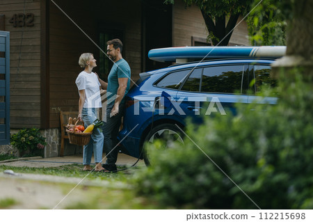Couple standing by their electric car. Electric vehicle with charger in charging port. Couple standing by their electric car. Electric vehicle with charger in charging port. 112215698