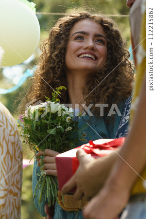 Portrait of surprised birthday girl at garden birthday party. Friends holding birthday gifts and flowers. 112215862