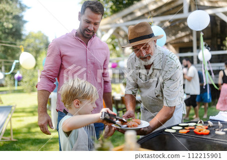 Father, grandfather and son grilling together at garden BBQ. Three generations of men at summer family garden party. 112215901