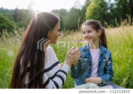 Beautiful mother with daughter, holding hands, sitting in the grass at meadow. Concept of Mother's Day and maternal love. 112215933