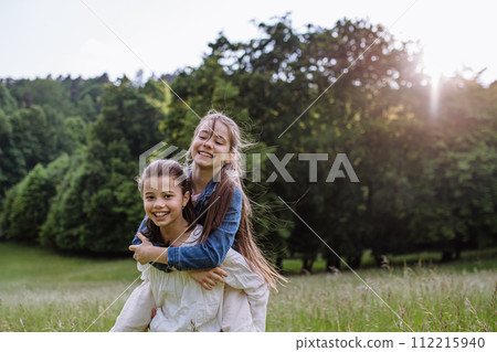 Two sisters playing at meadow in grass, having fun, piggybacking each other. Sisterly love and siblings relationship concept. Two sisters playing at meadow in grass, having fun, piggybacking each other. Sisterly love and siblings relationship concept. 112215940