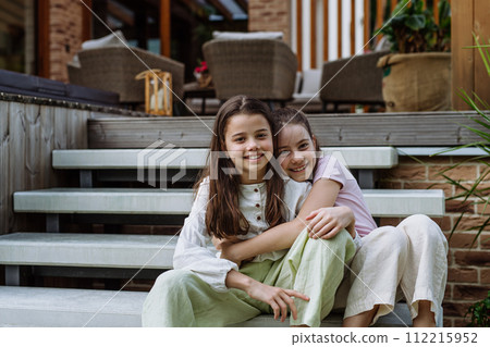 Two sisters sitting on stairs in front of house, embracing. Sisterly love and siblings relationship concept. 112215952