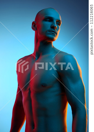 Portrait of handsome, serious bald young man with muscular, sportive body posing shirtless against blue studio background in neon light Portrait of handsome, serious bald young man with muscular, sportive body posing shirtless against blue studio background in neon light 112216065