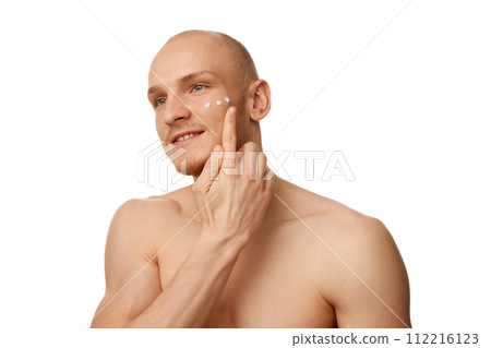 Handsome bald shirtless young man with muscular body, applying moisturizing face cream isolated against white studio background 112216123