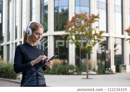 A focused woman enjoying her playlist while handling her smartphone in a leisurely stance, dressed in fitness attire, with modern architecture and greenery in the background. A focused woman enjoying her playlist while handling her smartphone in a leisurely stance, dressed in fitness attire, with modern architecture and greenery in the background. 112216329