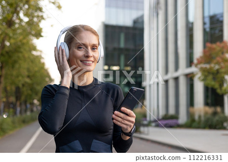 A young adult woman wearing athletic gear listens to music on her wireless headphones while holding a smartphone, evoking a sense of leisure and technology in an urban setting. 112216331