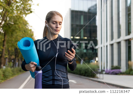 A focused female athlete carrying a yoga mat and hydration, listening to music or a podcast, and checking her smartphone before an outdoor fitness session in a city environment. 112216347