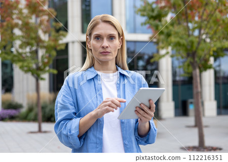 A focused professional woman stands outside on a cloudy day, using a digital tablet in front of a contemporary office building, embodying modern business and technology. A focused professional woman stands outside on a cloudy day, using a digital tablet in front of a contemporary office building, embodying modern business and technology. 112216351