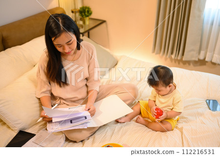 Beautiful business mom working with documents on on bed with her baby boy at home Beautiful business mom working with documents on on bed with her baby boy at home 112216531