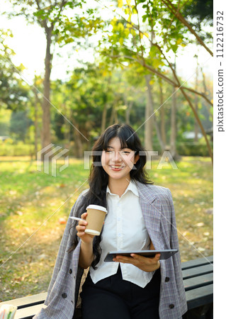 Portrait of smiling businesswoman in stylish suit using digital tablet on park bench 112216732