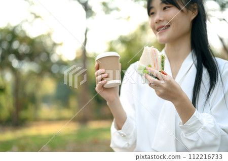 Smiling female employee having lunch break with coffee and sandwich while sitting on bench in the park 112216733