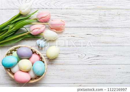 Happy Easter composition. Easter eggs in basket on colored table with yellow Tulips. Natural dyed colorful eggs background top view with copy space 112216997