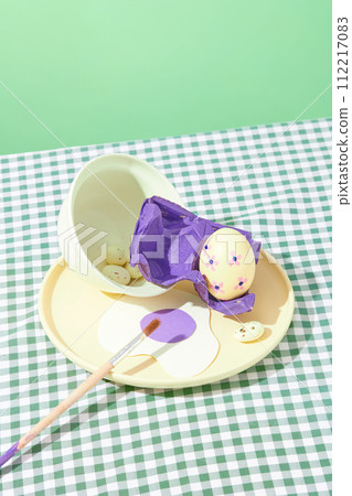 On a pastel green background and checkered plain, a round dish with Easter eggs, brush and purple blister decorated. Easter celebration concept On a pastel green background and checkered plain, a round dish with Easter eggs, brush and purple blister decorated. Easter celebration concept 112217083
