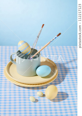 Easter eggs hand painted with ceramic dishes, cup and paintbrush decorated on light blue background. Egg decoration ideas for Easter, advertising photo 112217123