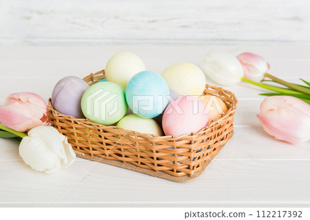Happy Easter composition. Easter eggs in basket on colored table with yellow Tulips. Natural dyed colorful eggs background with copy space Happy Easter composition. Easter eggs in basket on colored table with yellow Tulips. Natural dyed colorful eggs background with copy space 112217392