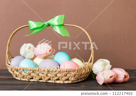 Happy Easter composition. Easter eggs in basket on colored table with yellow Tulips. Natural dyed colorful eggs background with copy space 112217402