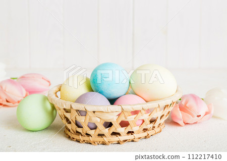 Happy Easter composition. Easter eggs in basket on colored table with yellow Tulips. Natural dyed colorful eggs background with copy space Happy Easter composition. Easter eggs in basket on colored table with yellow Tulips. Natural dyed colorful eggs background with copy space 112217410