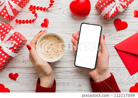 Female hands holding smart phone with coffee for Valentine day, gift box and envelope, hearts 112217899