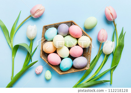 Happy Easter composition. Easter eggs in basket on colored table with yellow Tulips. Natural dyed colorful eggs background top view with copy space 112218128